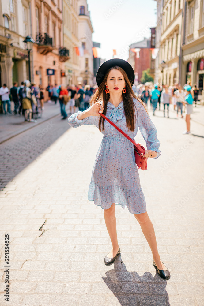 Young cheerful brunette woman in elegant dress and hat posing on city streer with people on background