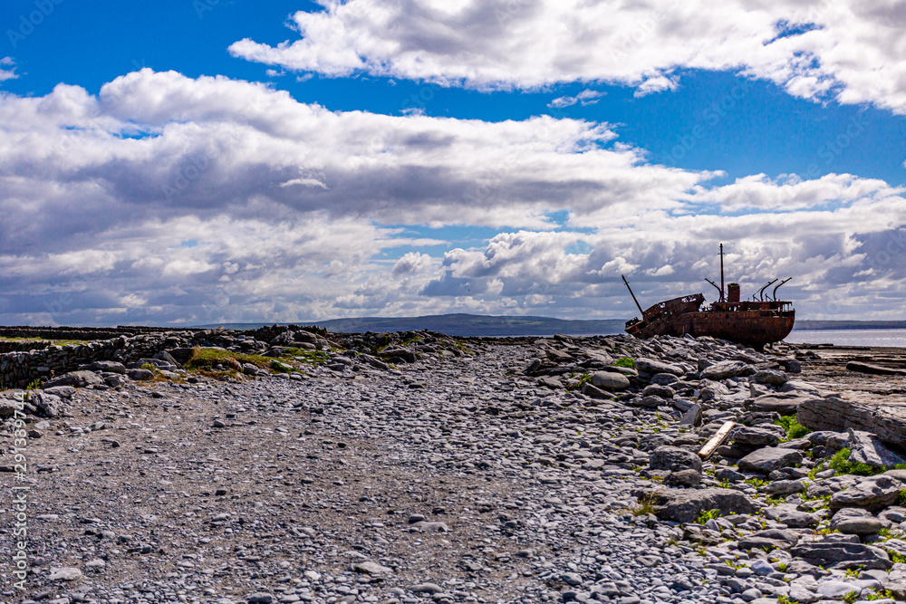 View of a stone footpath on the island of Inis Oirr with the shipwreck of Plassey in the background, abandoned ship, old and rusty over time, wonderful sunny day in the Aran Islands, Ireland