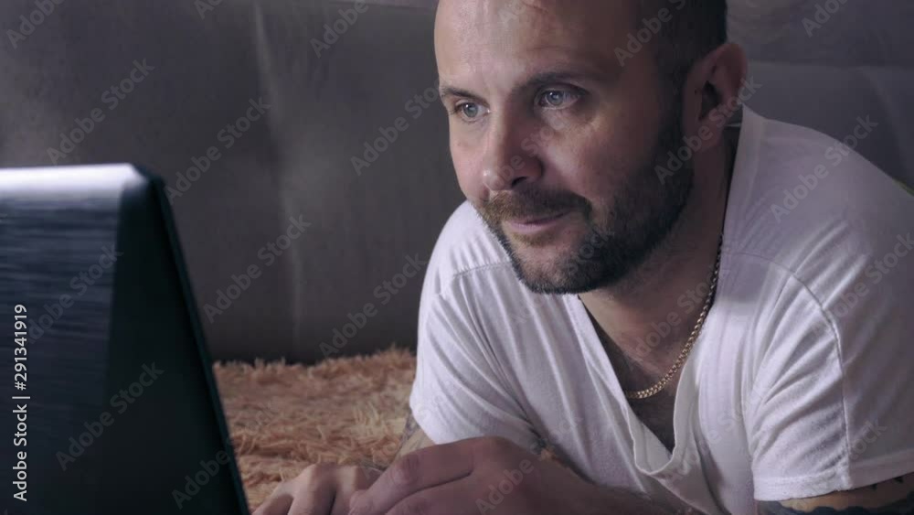 A man lies on a sofa and plays on a laptop. Night time. A man lies on a sofa and plays on a laptop. Night time.