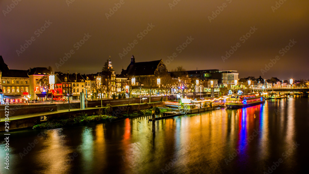 Night cityscape of the river Maas with its promenade, pier with a boat anchored with buildings, illuminated by multi-colored lights and reflected on the water, Maastricht, South Limburg, Netherlands
