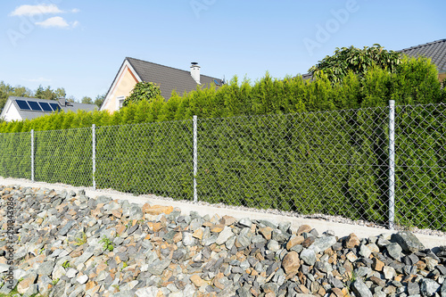 Street fence made of steel mesh, concrete is installed, pebble stones are below. Thuja green hedge grows next to the fence.