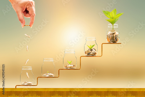 Investor hand hold and drop a gold coin in the five bottle and plant growing on golden steps on sunlight background, Business investment and saving money concept.