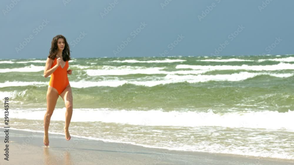 Brunette young woman in orange swimwear is running along the sandy beach, while waves touch her feet. Brunette young woman in orange swimwear is running along the sandy beach, while waves touch her feet.