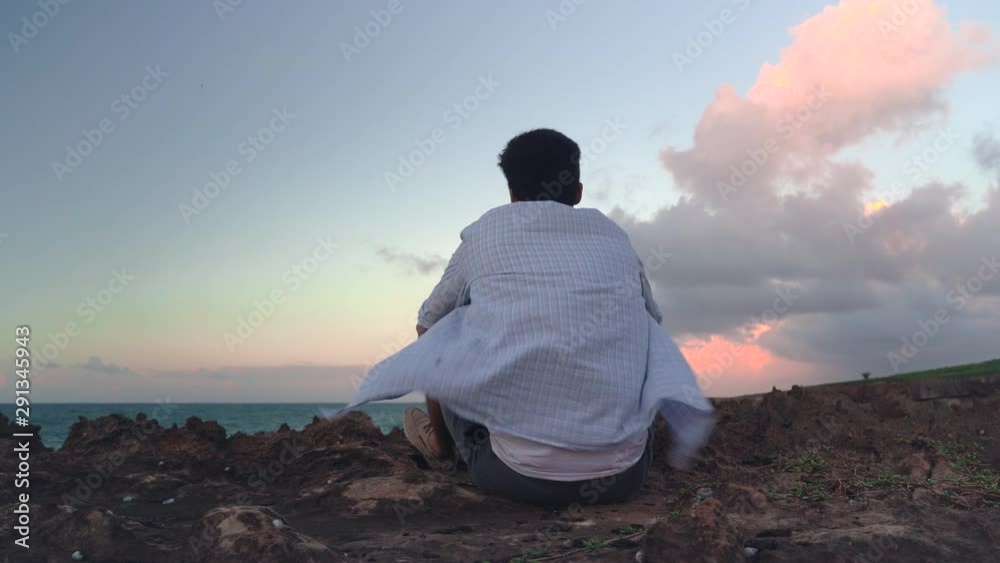 A medium angled shot of a depressed young man looking out to the horizon as he sits on volcanic rock in Puerto Rico. The sea stretches out in front, the pink breezy sunset bruises the sky above. A medium angled shot of a depressed young man looking out to the horizon as he sits on volcanic rock in Puerto Rico. The sea stretches out in front, the pink breezy sunset bruises the sky above.