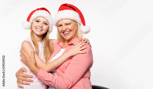 Beautiful senior grand mother in a pink shirt and Santa hat with light shoulder-length hair hugs her teenage blonde grand daughter in Christmas hat and laughs with her while looking at the camera.