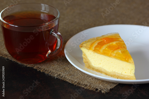 Cheesecake with oranges on a white plate, glass mug with tea on burlap tablecloth.