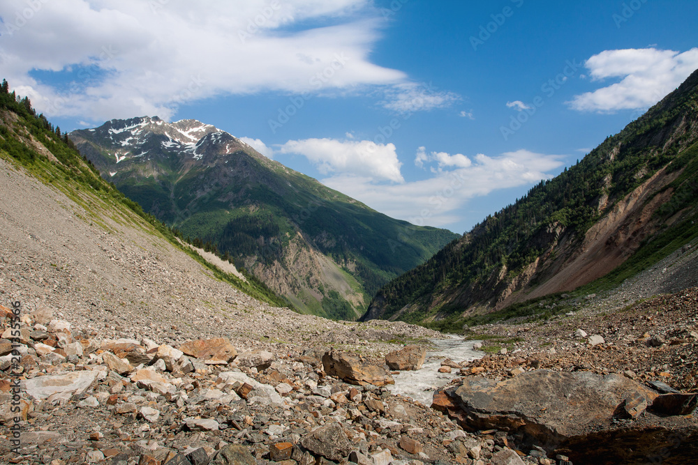 Mountain river flows through the valley. Stones lie on the banks of the river. In the background rise a mountain with a snowy peak. Mountain river flows through the valley. Stones lie on the banks of the river. In the background rise a mountain with a snowy peak.