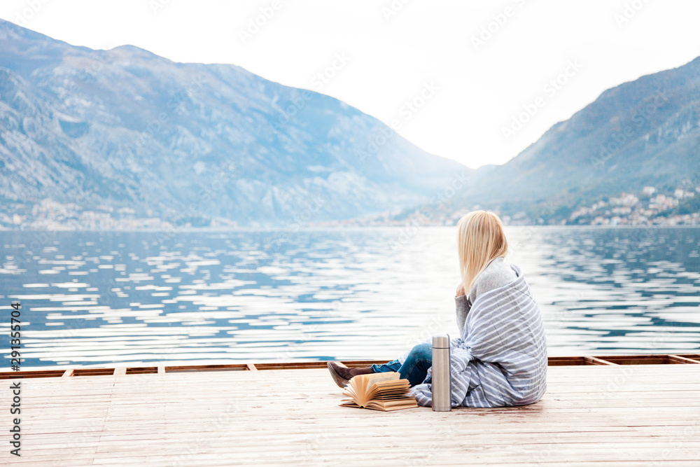 Fototapeta premium Woman on wooden pier by winter sea, mountains. Cozy picnic with coffee, hot beverages, tea in thermos and mug, warm plaid, opened book. Girl is enjoying nature, relaxation, reading on beach