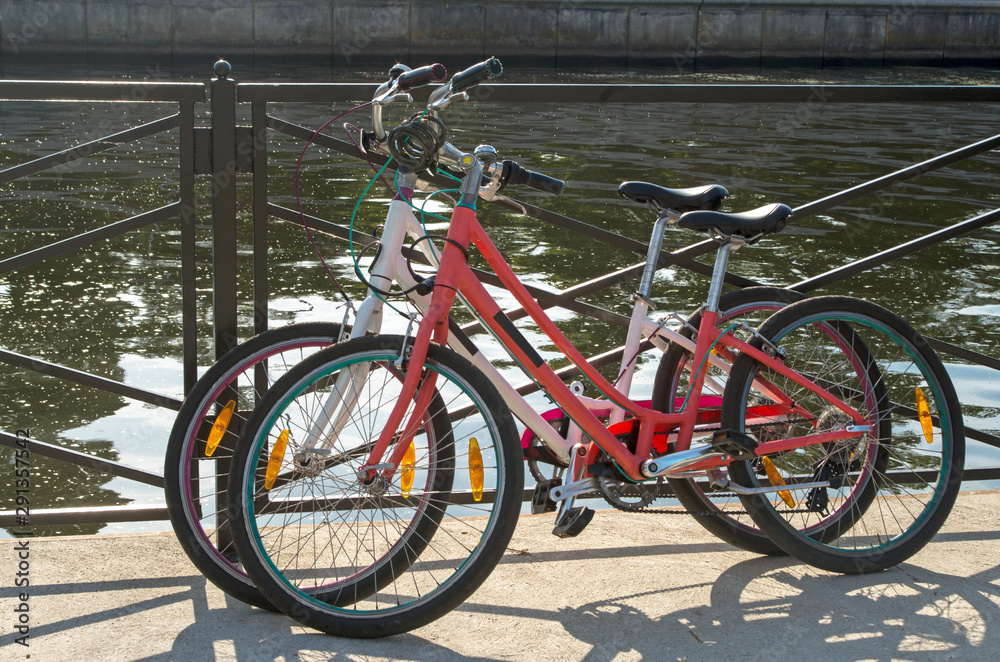 Fototapeta premium In the midday two bikes white and red of travelling together persons parked and locked with anti-theft cables to the fence street on the background of azure water in the river