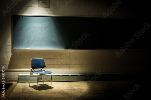 An Empty School Chair in a Dark, Shadowy Classroom - in Front of a Chalkboard with a Single Beam of Light Overhead