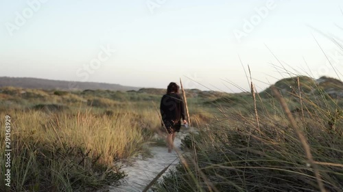 Woman walking in a sandy path surrounded by green and yellow foliage.
