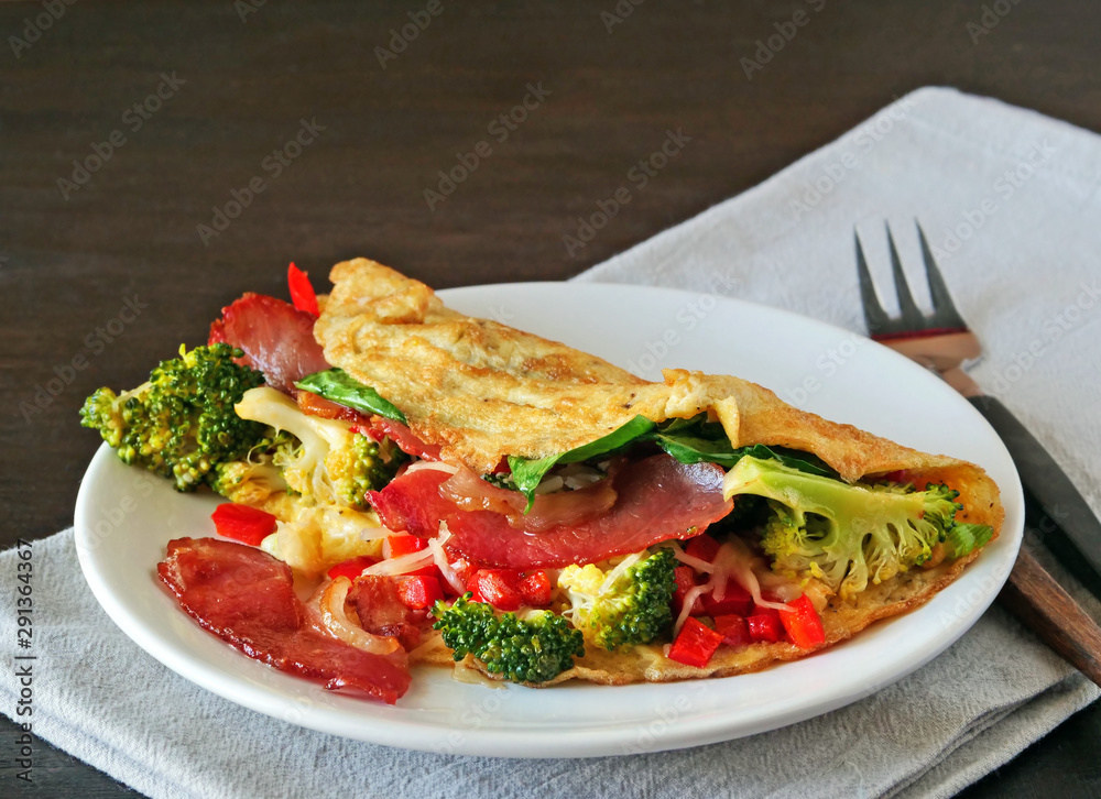 Tasty omelette with bacon, cheese, spinach and broccoli on white plate over dark background. Ketogenic folded omlette stuffed with low-carb high fat ingredients.