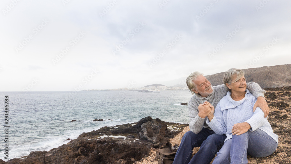 Two romantic people. Senior couple with grey hair sitting on the cliff looking away. Waiting for the sunrise in front to the ocean.