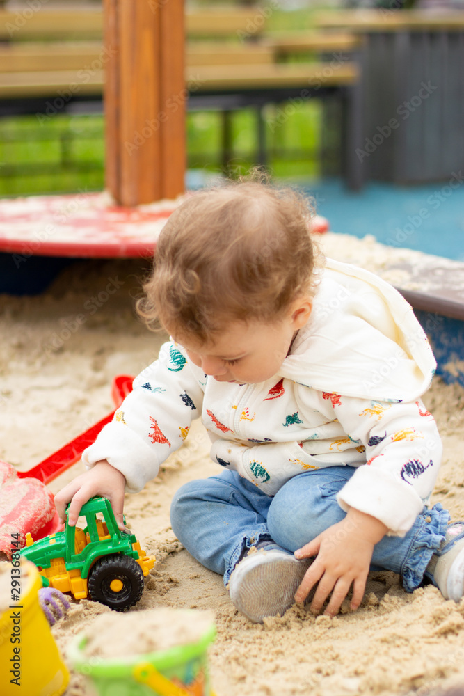 Fototapeta premium Baby girl boy in a white hoodie and jeans plays with a toy tractor. Happy Caucasian child 1 year old on a walk, vertical portrait