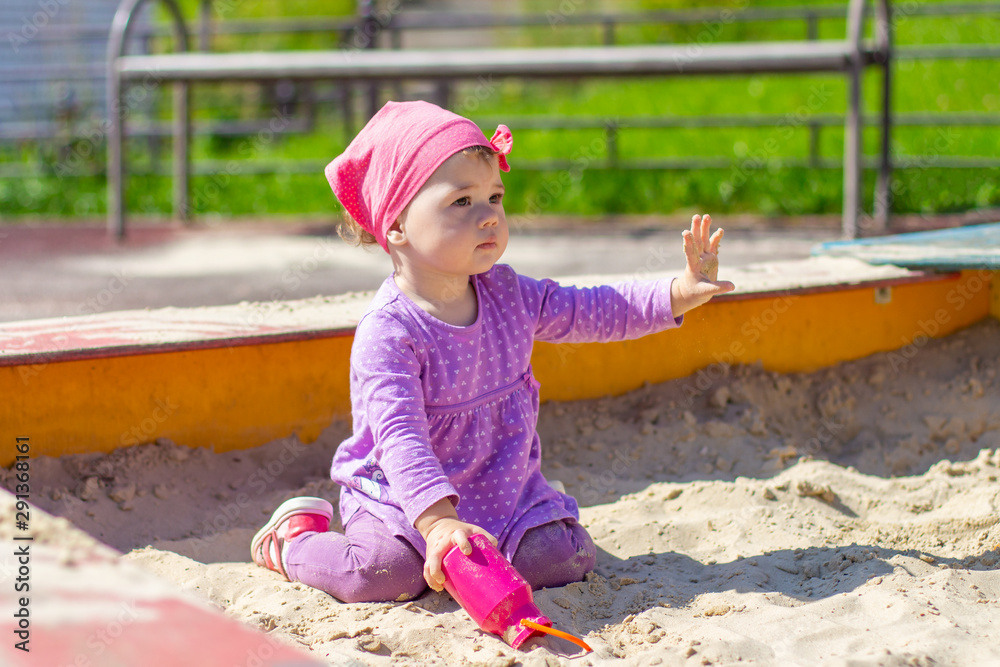 Baby girl in a purple dress plays with a toy pink jug outdoor. Happy Caucasian child 1 year old walking in the sandbox with raised hand Baby girl in a purple dress plays with a toy pink jug outdoor. Happy Caucasian child 1 year old walking in the sandbox with raised hand