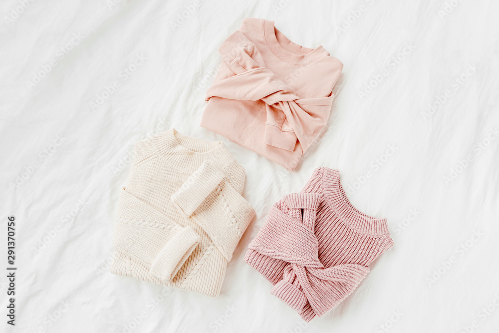 Three pale pink and white warm sweaters on bed. Women's stylish autumn or winter clothes. Cozy Winter look.  Flat lay, top view.