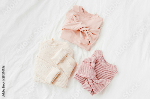 Three pale pink and white warm sweaters on bed. Women's stylish autumn or winter clothes. Cozy Winter look. Flat lay, top view.