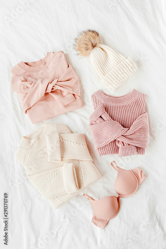 Three pale pink and white warm sweaters and winter hat on bed. Women's stylish clothes. Cozy Winter look. Flat lay, top view.