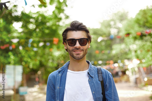 Goog looking young dark haired male with beard wearing casual clothes and sunglasses, standing over green park on sunny warm day, positive emotions concept