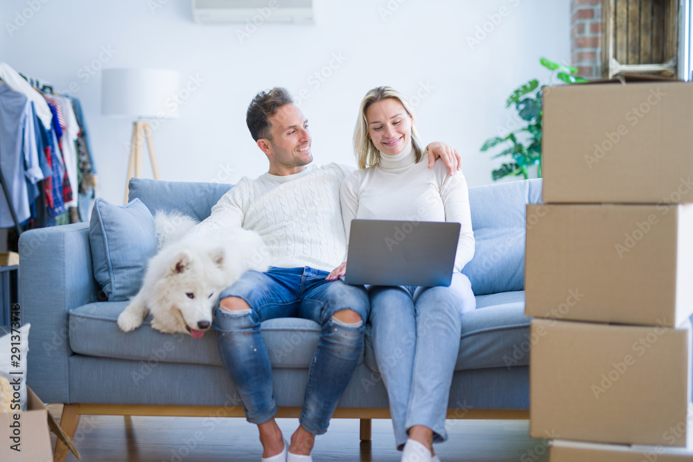 Young beautiful couple with dog sitting on the sofa using laptop at new home around cardboard boxes Young beautiful couple with dog sitting on the sofa using laptop at new home around cardboard boxes