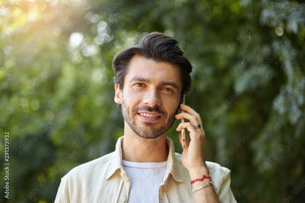 Outdoor close-up of charming young dark haired man with beard having talk on his mobile phone while walking along green garden on sunny day Outdoor close-up of charming young dark haired man with beard having talk on his mobile phone while walking along green garden on sunny day