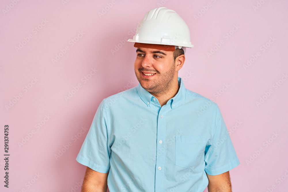 Young architect man wearing security helmet standing over isolated pink background looking away to side with smile on face, natural expression. Laughing confident.