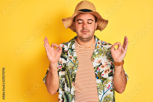 Young man on vacation wearing summer shirt and hat over isolated yellow background relax and smiling with eyes closed doing meditation gesture with fingers. Yoga concept.