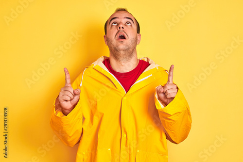 Young man wearing rain coat standing over isolated yellow background amazed and surprised looking up and pointing with fingers and raised arms.