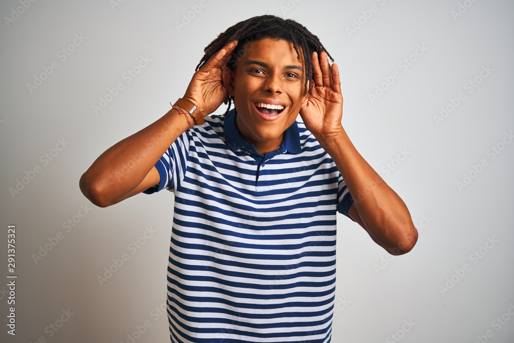 Afro man with dreadlocks wearing striped blue polo standing over isolated white background Smiling pulling ears with fingers, funny gesture. Audition problem