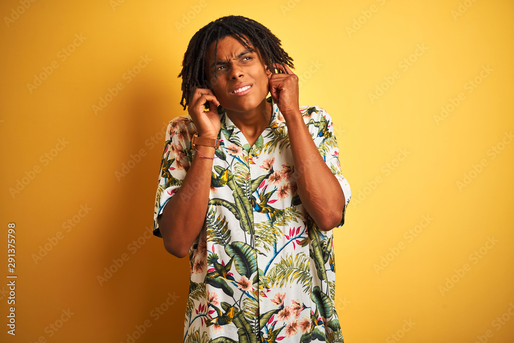 Afro man with dreadlocks on vacation wearing summer shirt over isolated yellow background covering ears with fingers with annoyed expression for the noise of loud music. Deaf concept. Afro man with dreadlocks on vacation wearing summer shirt over isolated yellow background covering ears with fingers with annoyed expression for the noise of loud music. Deaf concept.