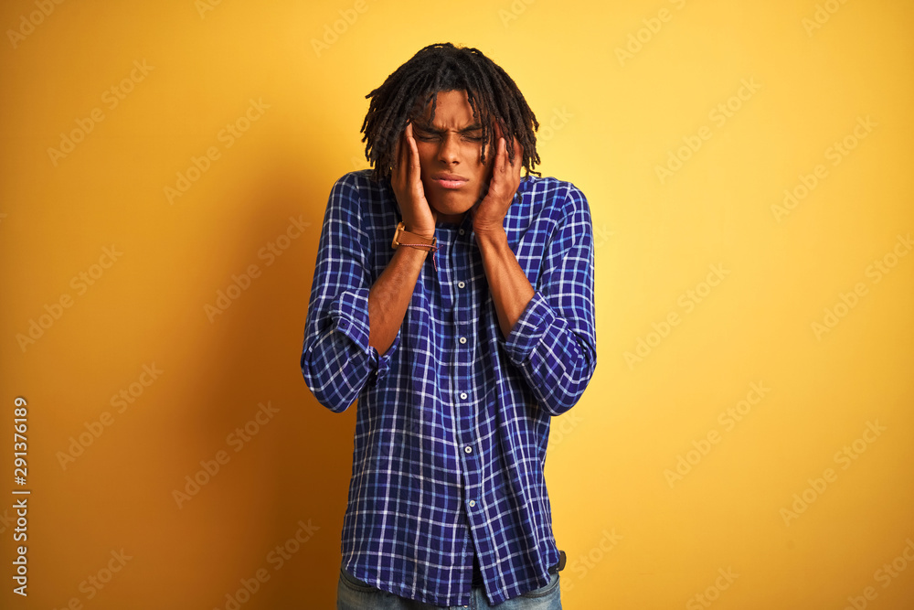 Afro man with dreadlocks wearing casual shirt standing over isolated yellow background with hand on head for pain in head because stress. Suffering migraine. Afro man with dreadlocks wearing casual shirt standing over isolated yellow background with hand on head for pain in head because stress. Suffering migraine.