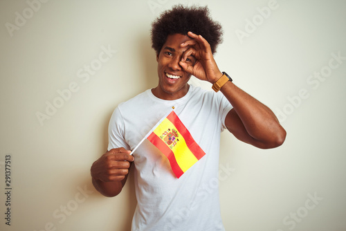 Young afro american man holding Spain Spanish flag standing over isolated white background with happy face smiling doing ok sign with hand on eye looking through fingers