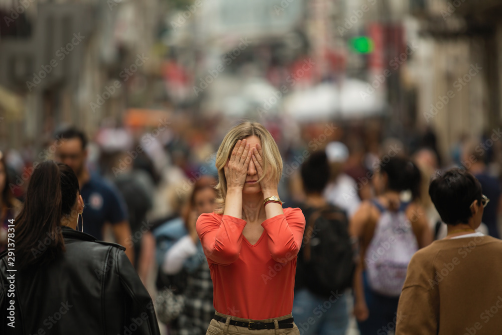 Sad depressed woman covers his eyes with his hands surrounded by people walking in crowded street. Panic attack in public place.
