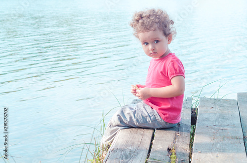 cute little pensive girl in the morning sits on a wooden pier on the shore of a forest lake, looks at the camera