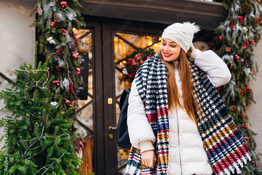 A girl in winter clothes and a backpack looks down and corrects a hat with a pompon near a beautiful cafe with glass doors and Christmas decorations.