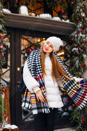 cute girl in winter clothes with a playful look and a smile near a beautiful cafe with glass doors and Christmas decorations.