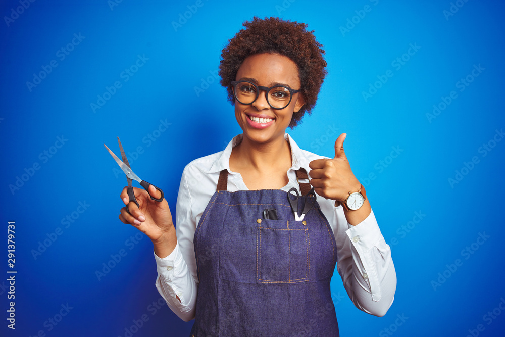 Fototapeta African american hairdresser woman holding scissors over blue isolated background happy with big smile doing ok sign, thumb up with fingers, excellent sign