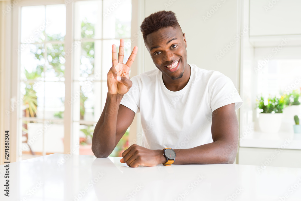 Handsome african american man on white table at home showing and pointing up with fingers number three while smiling confident and happy. Handsome african american man on white table at home showing and pointing up with fingers number three while smiling confident and happy.