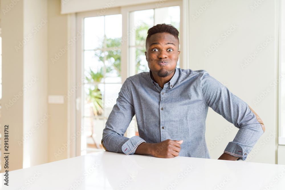 Handsome african american man on white table puffing cheeks with funny face. Mouth inflated with air, crazy expression.