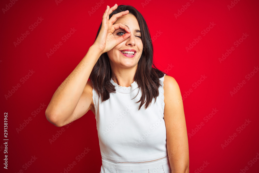 Young beautiful woman wearing white dress standing over red isolated background doing ok gesture with hand smiling, eye looking through fingers with happy face. Young beautiful woman wearing white dress standing over red isolated background doing ok gesture with hand smiling, eye looking through fingers with happy face.