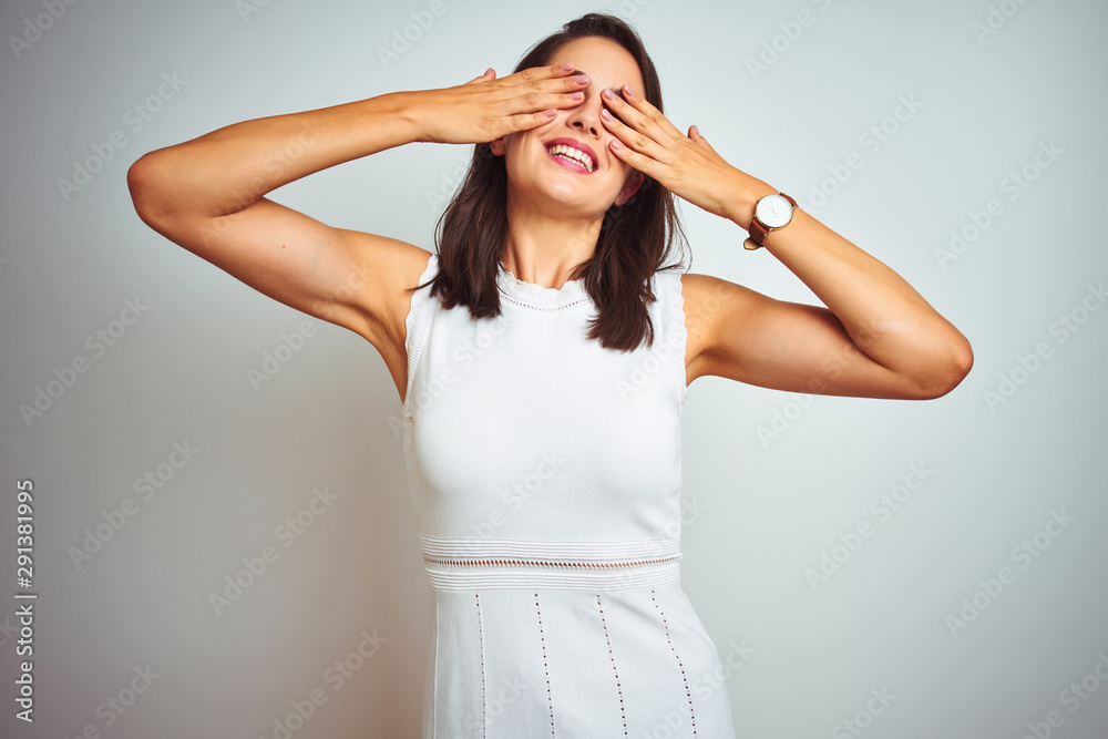 Young beautiful woman wearing dress standing over white isolated background covering eyes with hands smiling cheerful and funny. Blind concept.