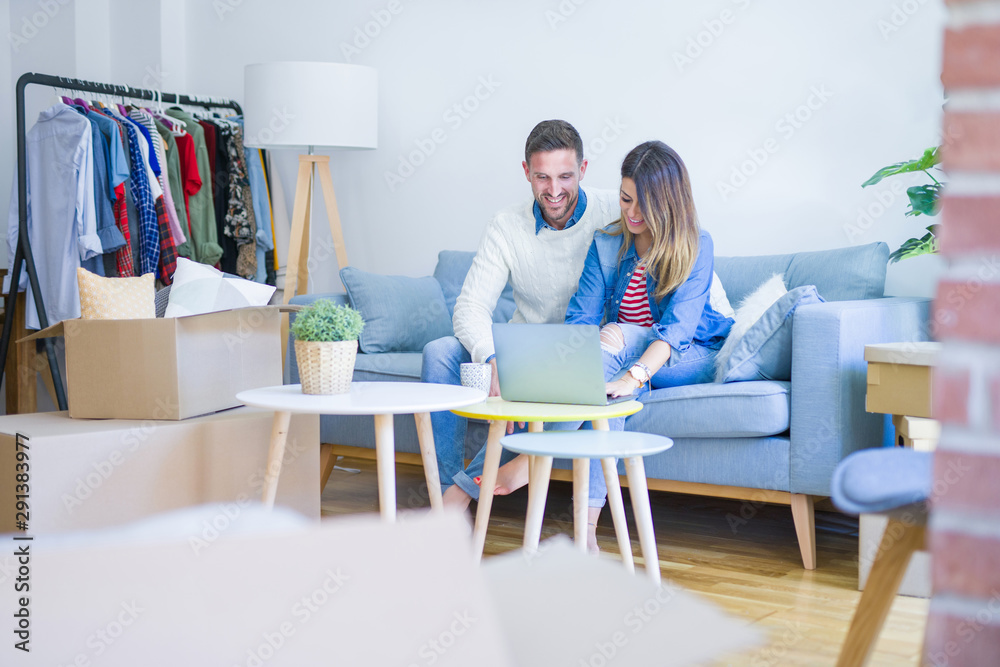 Young beautiful couple sitting on the sofa drinking cup of coffee using laptop at new home around cardboard boxes Young beautiful couple sitting on the sofa drinking cup of coffee using laptop at new home around cardboard boxes