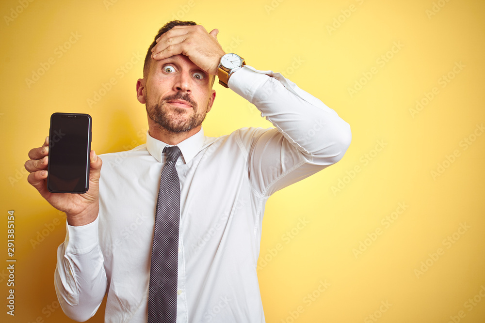 Young handsome business man showing smartphone screen over yellow isolated background stressed with hand on head, shocked with shame and surprise face, angry and frustrated
