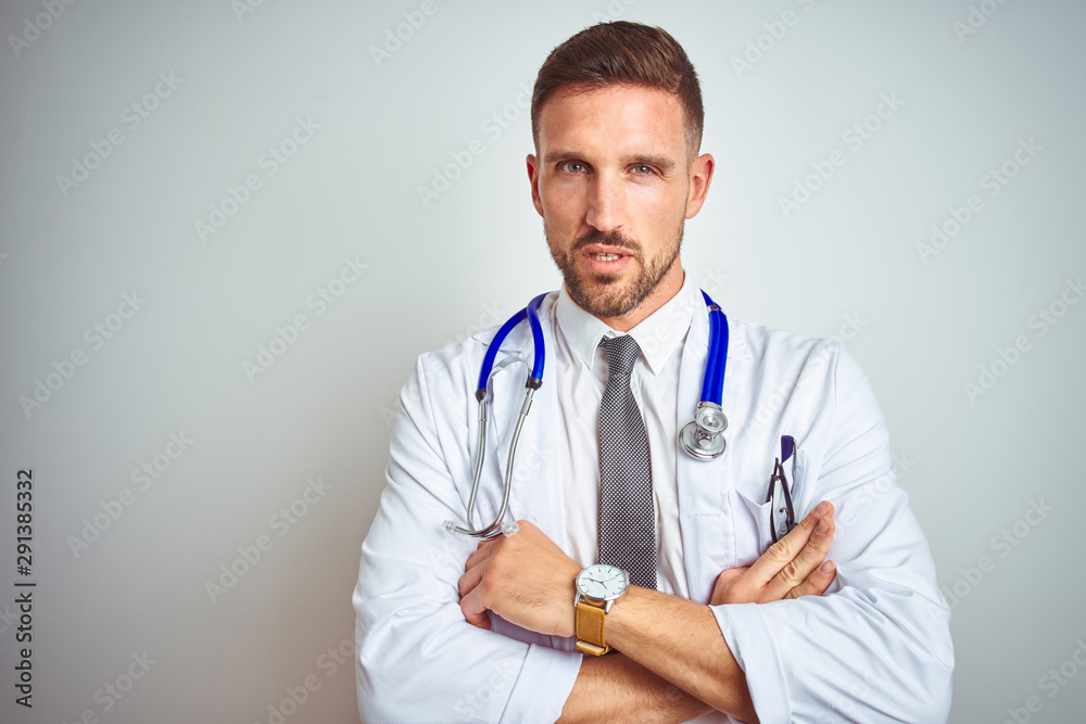 Young handsome doctor man wearing white professional coat over isolated background skeptic and nervous, disapproving expression on face with crossed arms. Negative person. Young handsome doctor man wearing white professional coat over isolated background skeptic and nervous, disapproving expression on face with crossed arms. Negative person.