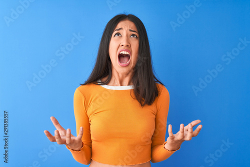 Young beautiful chinese woman wearing orange t-shirt standing over isolated blue background crazy and mad shouting and yelling with aggressive expression and arms raised. Frustration concept.