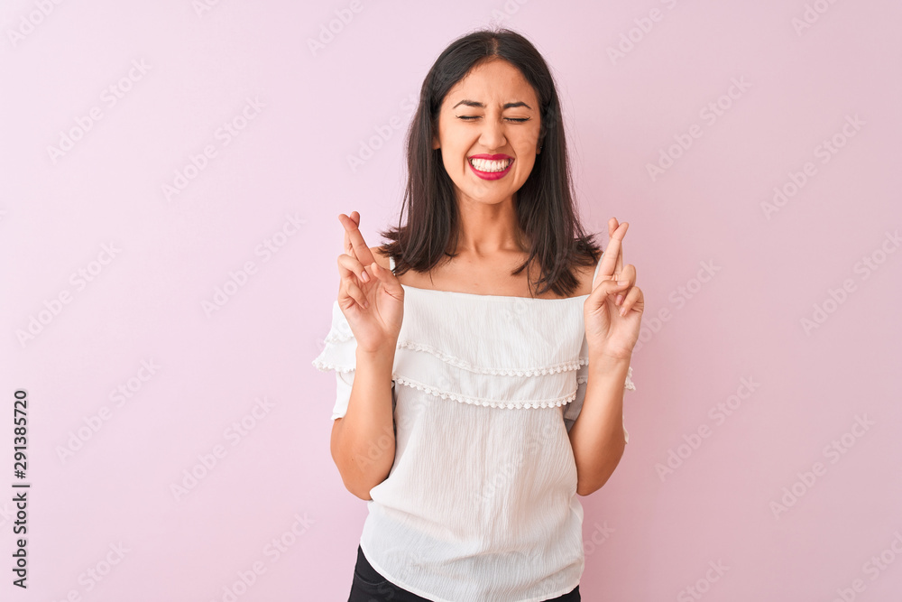 Beautiful chinese woman wearing white t-shirt standing over isolated pink background gesturing finger crossed smiling with hope and eyes closed. Luck and superstitious concept. Beautiful chinese woman wearing white t-shirt standing over isolated pink background gesturing finger crossed smiling with hope and eyes closed. Luck and superstitious concept.