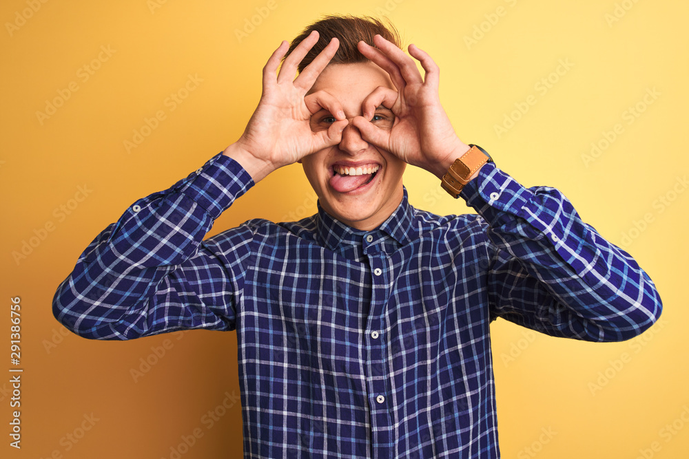 Young handsome man wearing casual shirt standing over isolated yellow background doing ok gesture like binoculars sticking tongue out, eyes looking through fingers. Crazy expression. Young handsome man wearing casual shirt standing over isolated yellow background doing ok gesture like binoculars sticking tongue out, eyes looking through fingers. Crazy expression.
