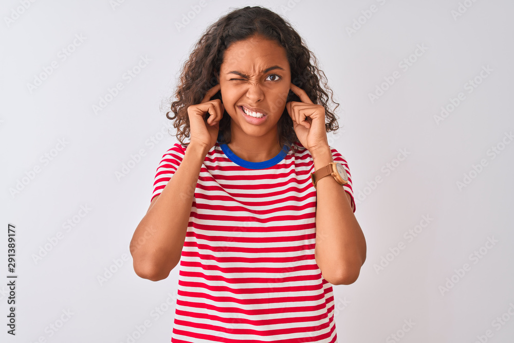 Young brazilian woman wearing red striped t-shirt standing over isolated white background covering ears with fingers with annoyed expression for the noise of loud music. Deaf concept. Young brazilian woman wearing red striped t-shirt standing over isolated white background covering ears with fingers with annoyed expression for the noise of loud music. Deaf concept.