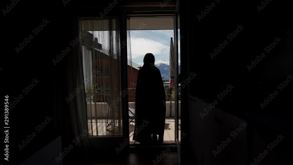 Young woman wrapped in a plaid go out to the terrace with scenic mountain view from the living room. Rear view