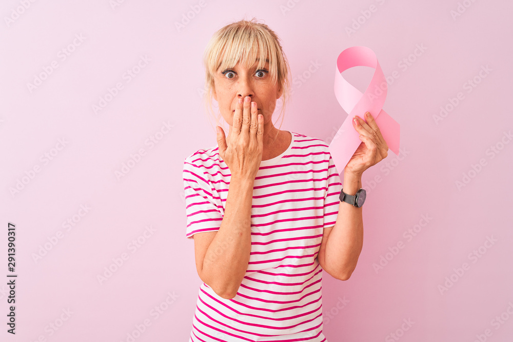 Middle age woman holding cancer ribbon standing over isolated pink background cover mouth with hand shocked with shame for mistake, expression of fear, scared in silence, secret concept Middle age woman holding cancer ribbon standing over isolated pink background cover mouth with hand shocked with shame for mistake, expression of fear, scared in silence, secret concept